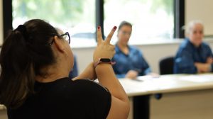 uma mulher branca em primeiro plano está fazendo um sinal com a mão direita onde todos os dedos da mão estão fechados exceto pelos dedos indicador e médio, fazendo um V com esses dois dedos. Ao fundo desfocado existem duas mulheres olhando para as mãos da mulher que faz o sinal em Libras.